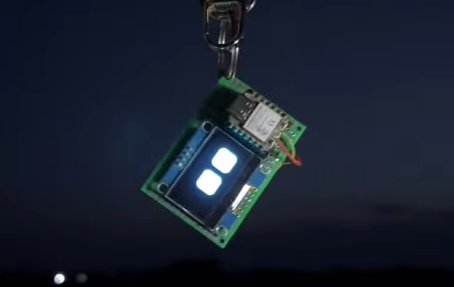 Small green circuit board with a compact display showing two glowing square icons, hanging from a metal keychain against a dark background.